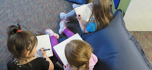 Three girls practice their letters on individual whiteboards.
