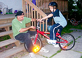 A father and son are on the back steps of their house. The father is making an adjustment to his young adolescent son’s bicycle.