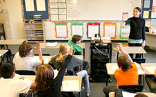 A smiling young female teacher is standing in front of her Middle Years class and pointing to a student who has her hand raised to answer a question.
